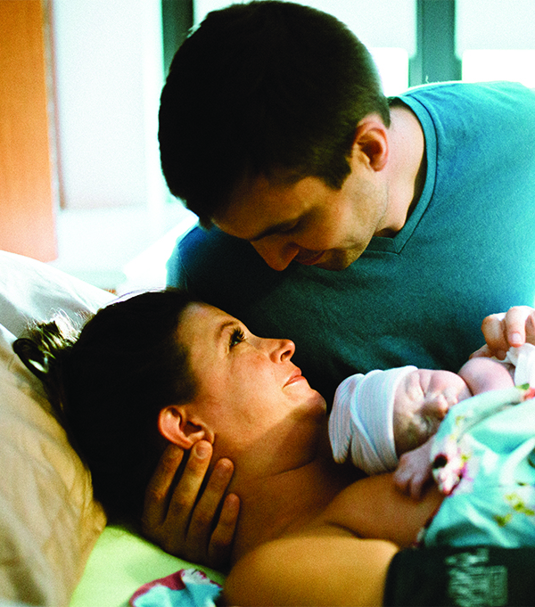 Photo by Michael Lathrop. Jesse, holding her newborn son Gus while smiling up at her husband.